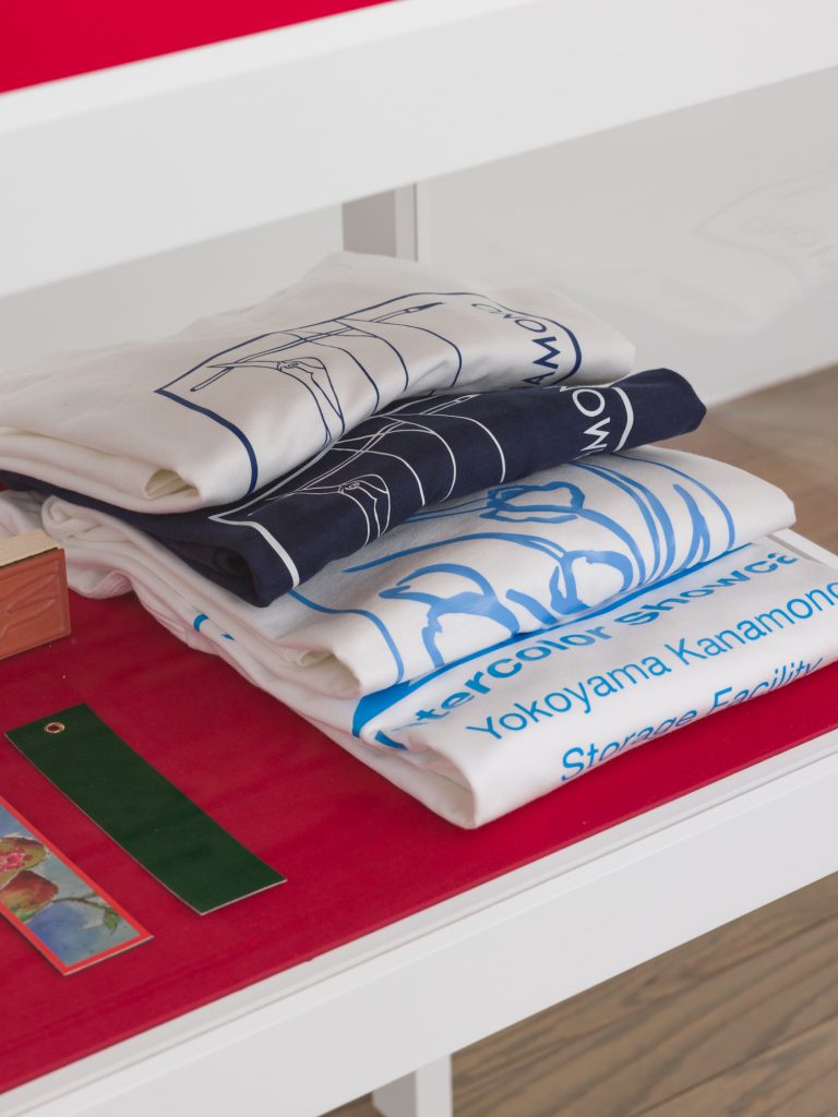 A detail image of Horibuchi's Showcase Vitrine 3 featuring four t-shirts that read "Watercolor Showcase Yokoyama Kanamono Storage Facility" folded atop red velvet. Image courtesy of Patron Gallery. Photographer Evan Jenkins.