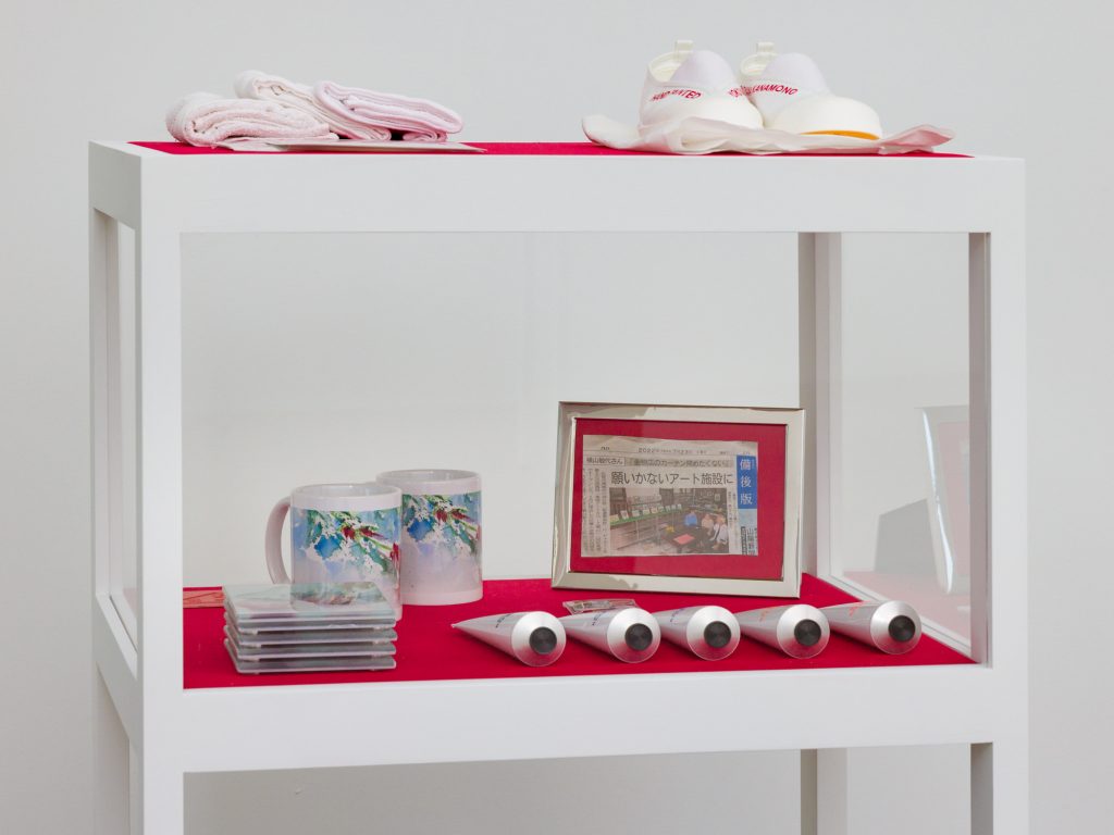 A two-tiered, white showcase vitrine with red velvet surfaces displays tubes of paint, branded mugs and coasters, and a pair of inscribed slip-on shoes. Image courtesy of Patron Gallery. Photographer Evan Jenkins.
