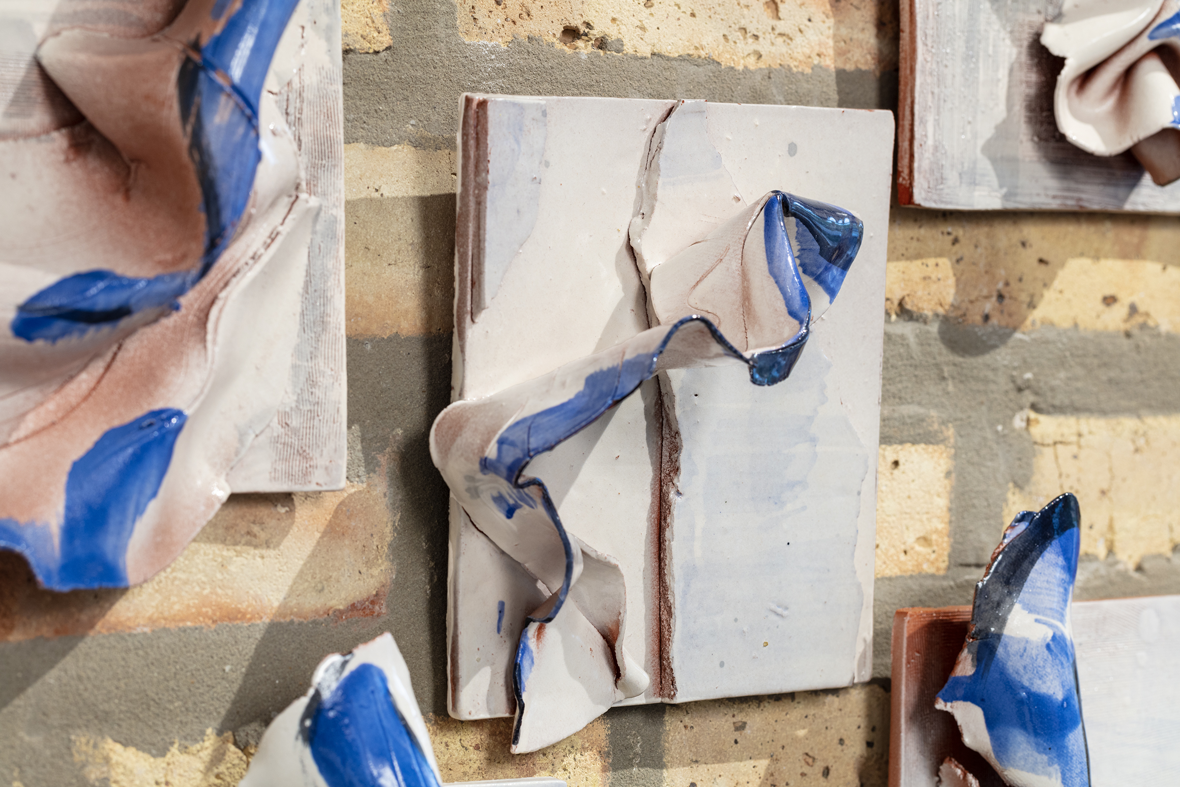 Image: On a brick wall, several of Nanxi Jin's sculptures hang. Each has a beige concrete square back and protruding beige elements, accented by a rich blue. Photo courtesy of the artist.