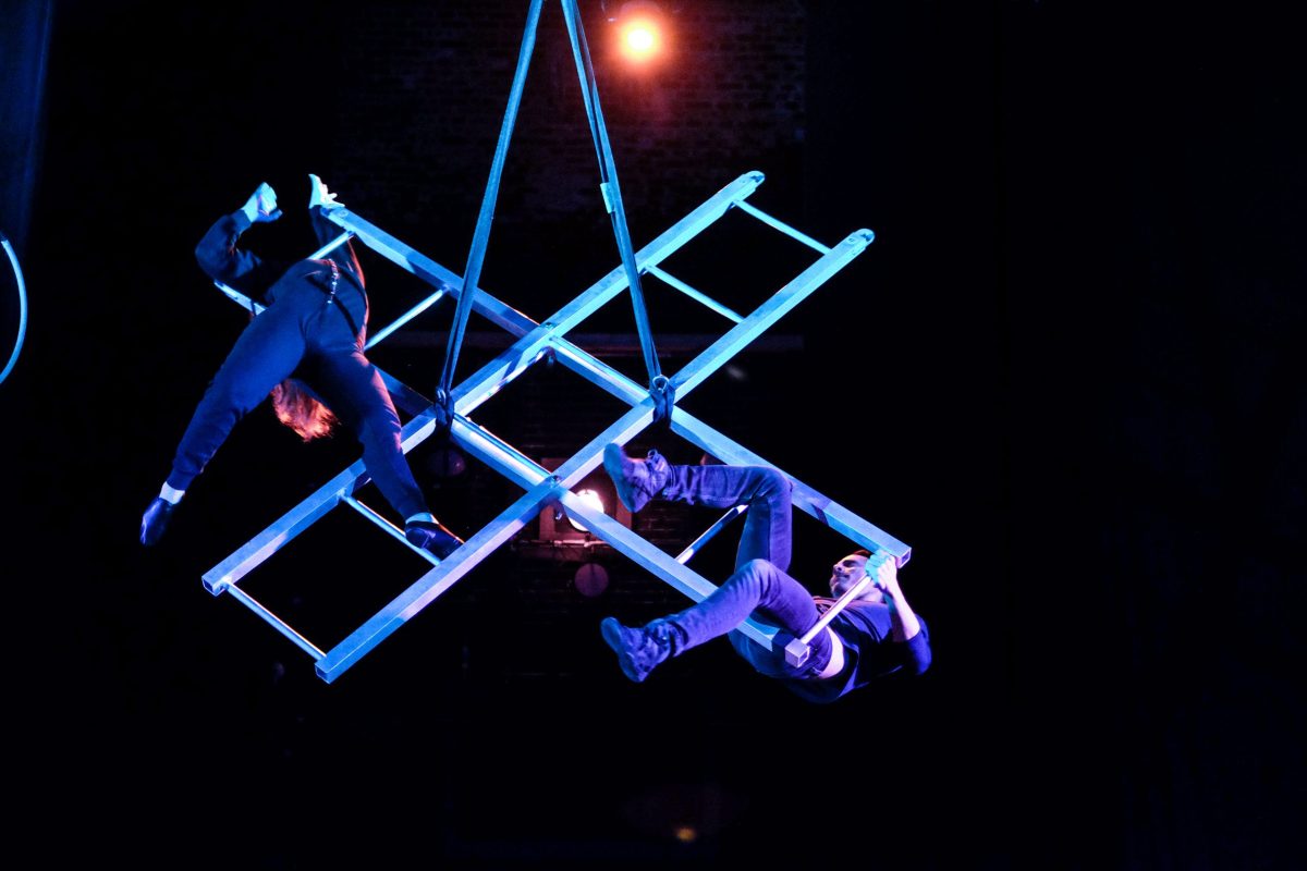Image: Performance at The Actors Gym. Bathed in blue light, two people are hanging from criss-crossed ladders hanging from the ceiling. One person is leaning. Photo by Matthew C. Yee.