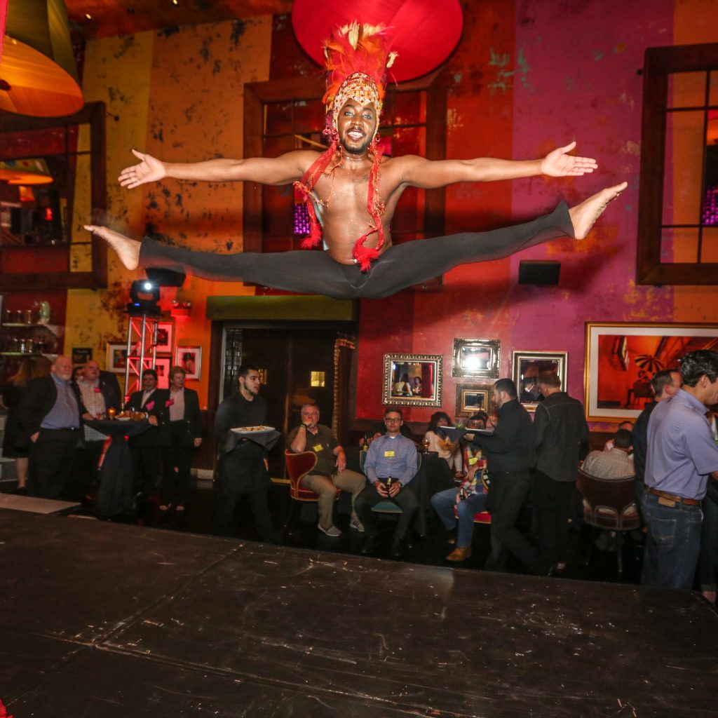 Image: An Actors Gym performer with a red and gold headdress leaps into the air on stage during a performance at a private event, while event-goers eat, drink, and look on. Photo by Will Byington Photography, courtesy of The Actors Gym.