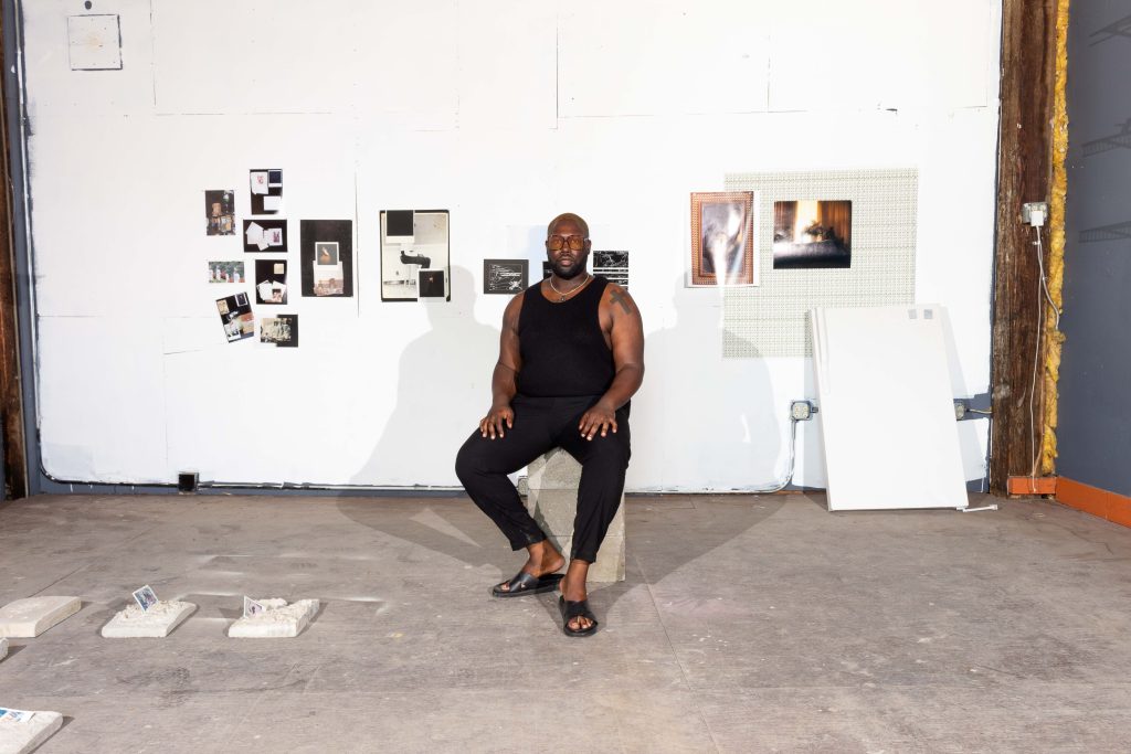Image: Shabez Jamal, Self-portrait in studio, 2023. A portrait of the artist in a studio space, surrounded by their artworks. Some photographic images hang just behind them with a refrigerator door propped up against the wall close by. At their feet and to the left are several concrete works from In Remembrance of Us. Image courtesy of Shabez Jamal.