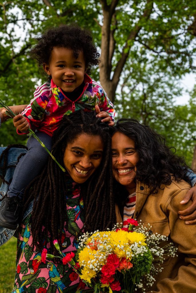 Image: Suyan sits on Taryn's shoulders as Vivi and Taryn huddle together in a close embrace. All three are smiling big at the camera, while Vivi holds a bouquet of flowers. Photo by Tonal Simmons.