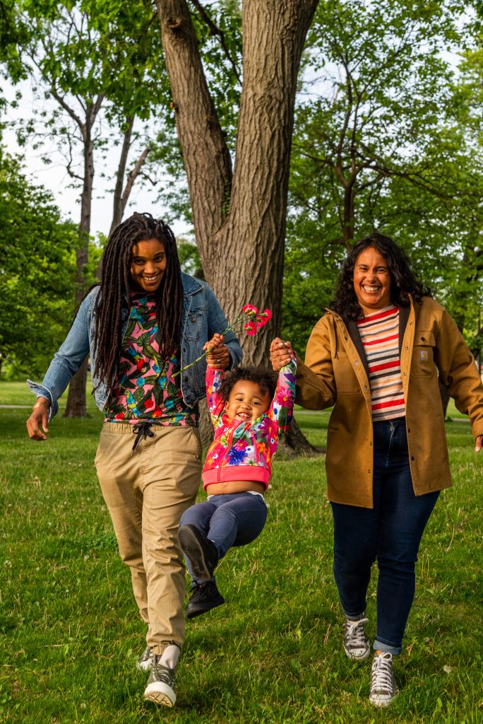 Image: Vivi and Taryn swing Suyan by their arms in between them. All three are smiling big and laughing. Photo by Tonal Simmons.
