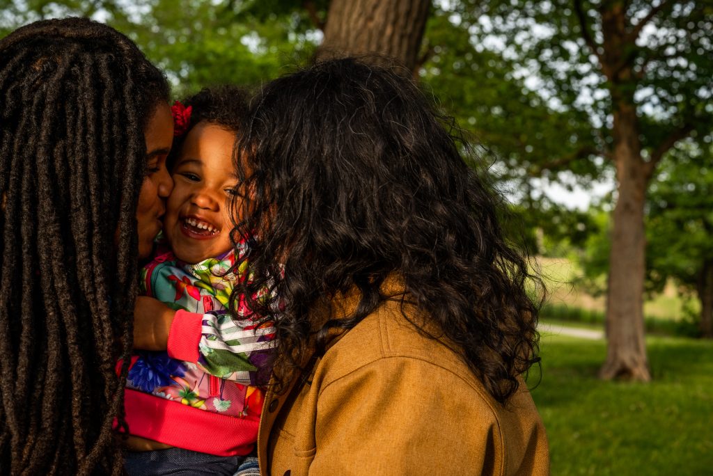 Taryn (left) and Vivi (right) kiss Suyan's cheeks as they hold her up between them. Suyan has a big grin on her face. Taryn's thick and long dreadlocks obscure part of their face while Vivi's hair obscures her face completely. Photo by Tonal Simmons.