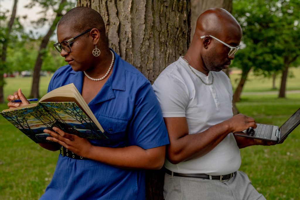 Image: Josey (left) and Johnny (right) are learning in front of a tree in Humboldt Park, turned away from each other with their arms touching each other. Josey is dressed in a blue dress and a black leather belt around her waist. She is wearing black glasses, a white pearl necklace and star earrings. She is holding a journal and a pen. Johnny is dressed in a gray short-sleeved henley shirt and gray slacks with a black leather belt. He wears white glasses and a silver necklace. He is looking down at a small laptop in his hand. Photo by Tonal Simmons.