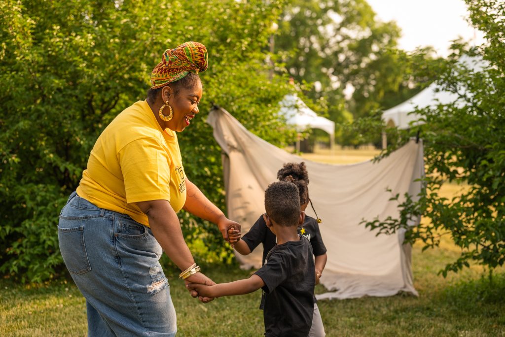CeeJaye, Hasan, and Naomi hold hands in a circle, laughing in a Chicago park.  Behind them is a white sheet that is clipped between two bushes and white tents. Photo by Tonal Simmons.