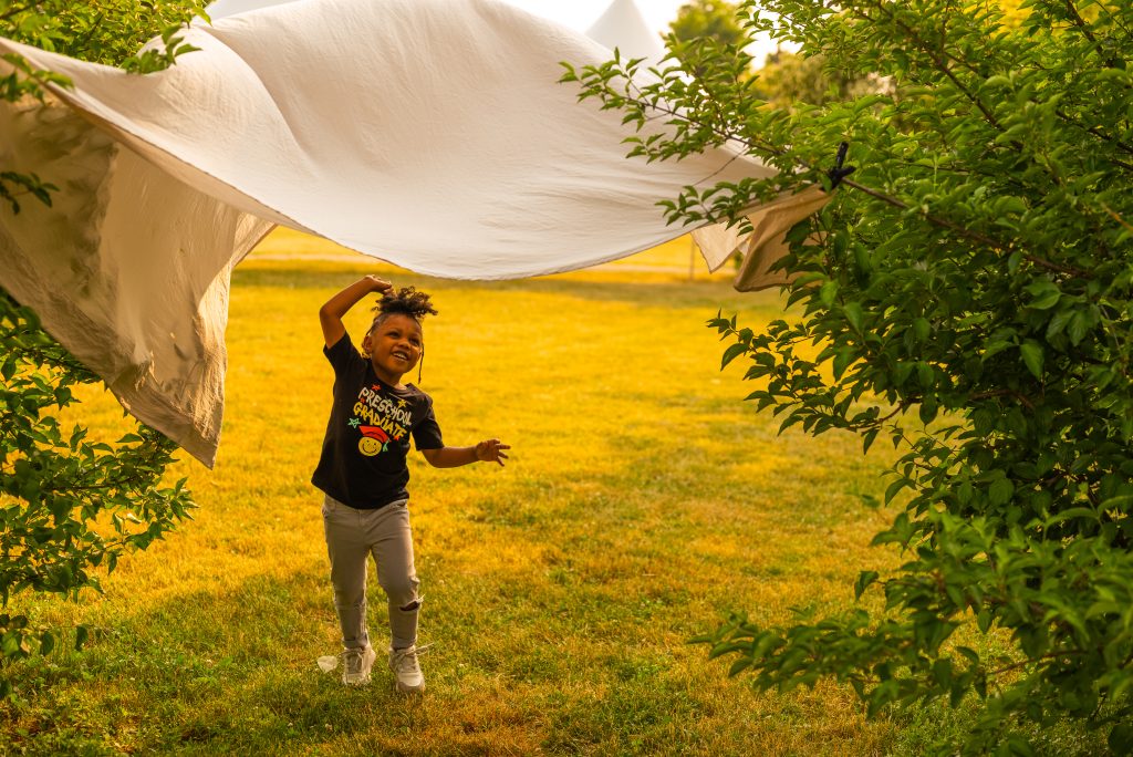 Image: Naomi smiles and plays with a white sheet that is tied between two bushes above her head. Photo by Tonal Simmons.