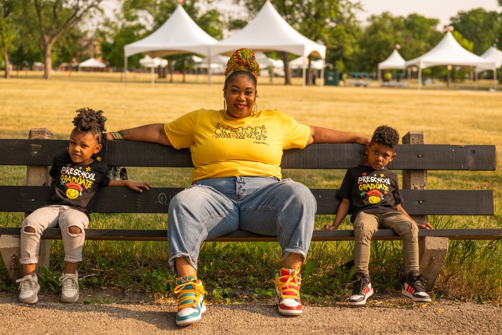 Image: CeeJaye, Hasan, and Naomi sit on a bench in in Humboldt Park. CeeJaye wears blue jeans, a bright yellow t-shirt, a multi-colored headwrap, and large hoop earrings. Hasan and Naomi wear black t-shirts and pants. CeeJaye, smiling, sits with both arms resting on the bench above Naomi and Hasan's head. Behind them, at a distance across a field of yellowed grass, are white tents. Photo by Tonal Simmons.