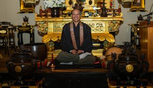 Image: Brown seated in front of a golden shrine. They smile broadly. Photo by Tonal Simmons.