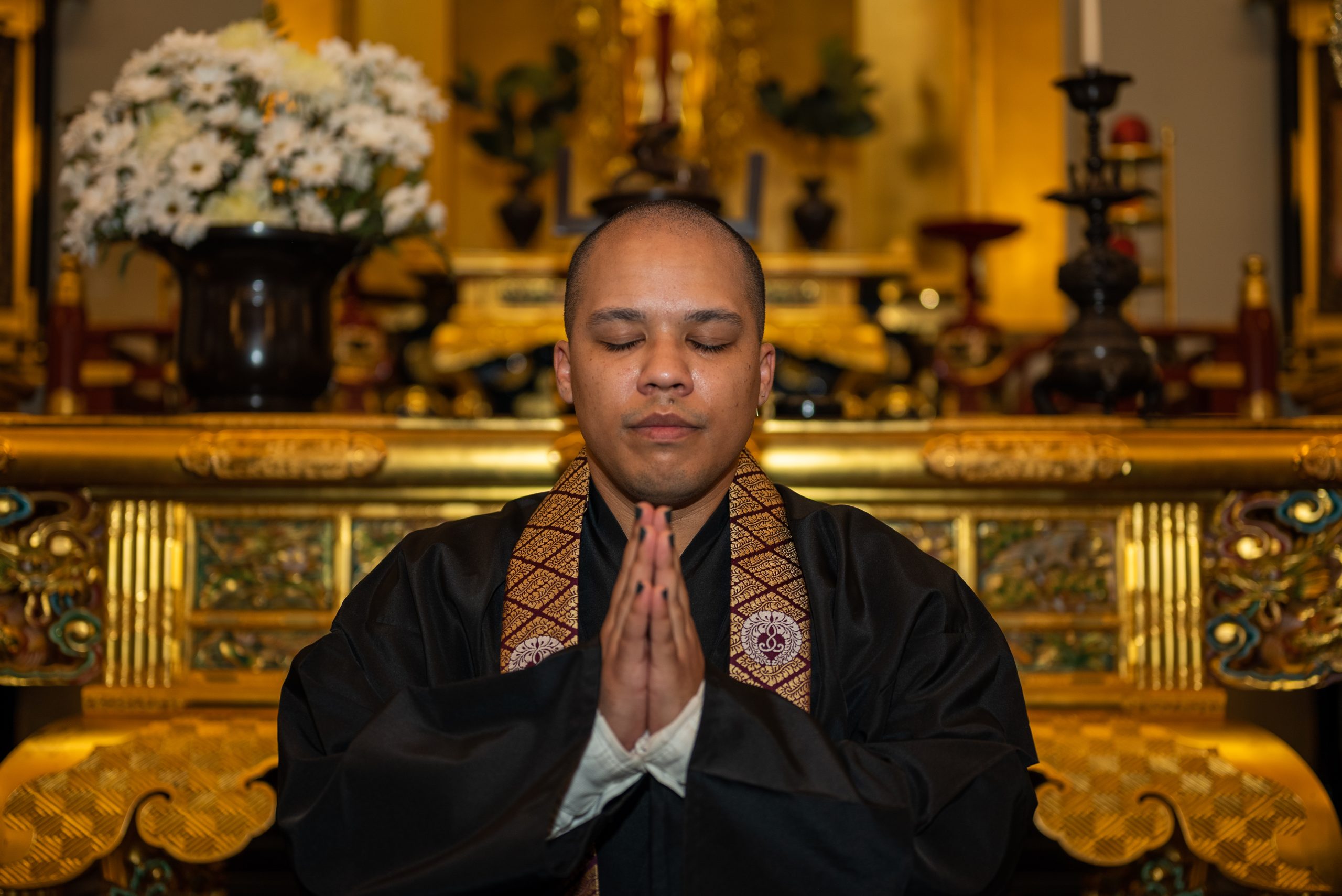 Image: Eli Ryn Brown, eyes closed, hands flat together. Behind them is a golden shrine with flowers and candles on it. Photo by Tonal Simmons.