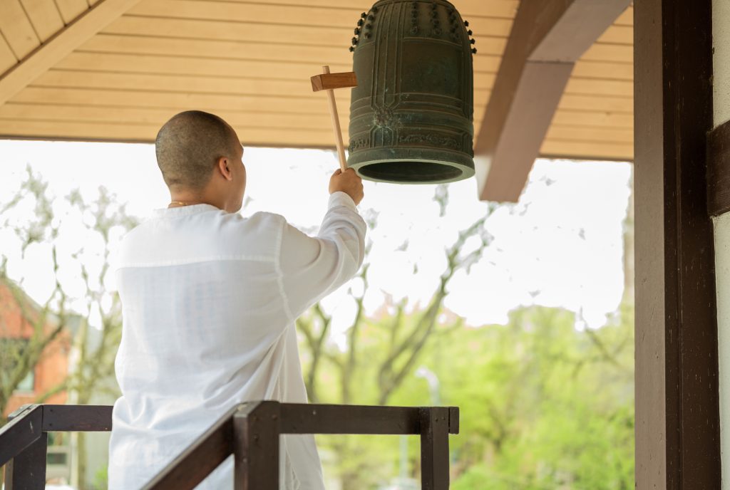 Image: Brown taps a large bell underneath an open outdoor structure. In the background, trees. Photo by Tonal Simmons.