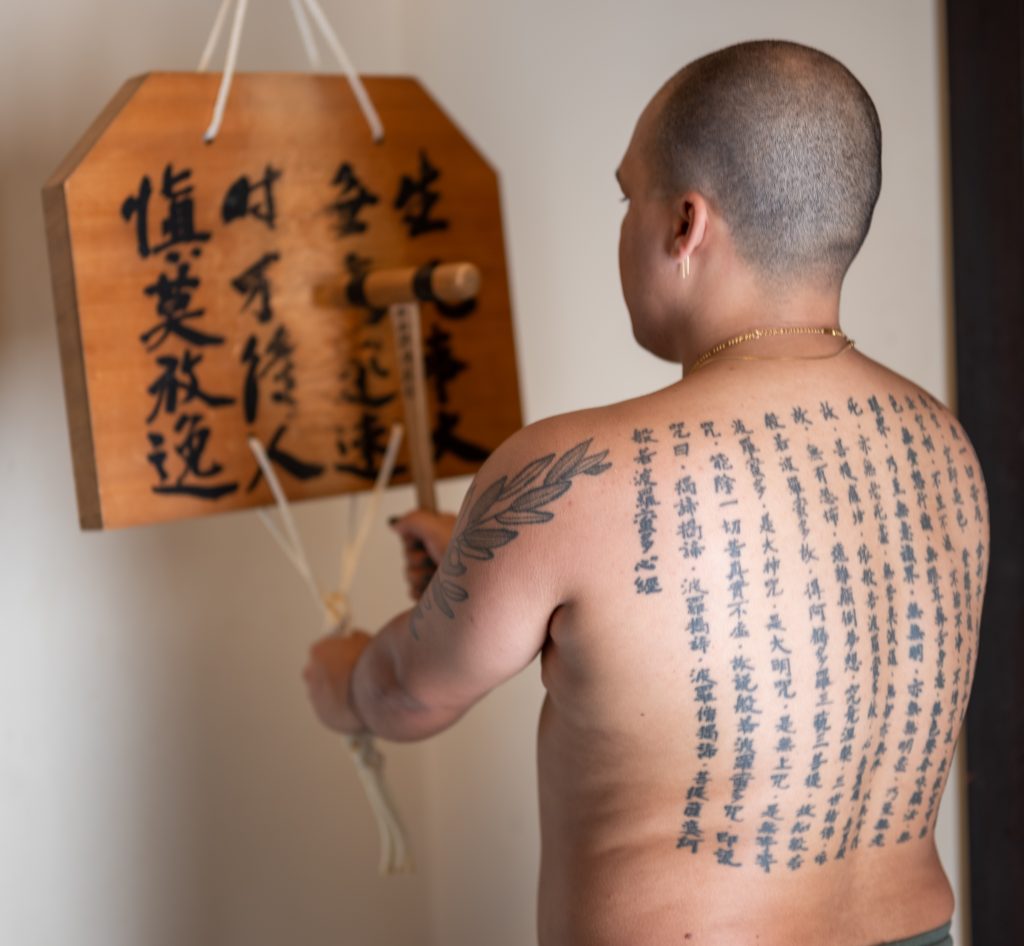 Image: Brown, facing away from us,  holding a wooden tool like a hammer. In their other hand, they hold ropes that are attached to the bottom of a heavy polished wooden plank with Japanese text written on it. On Brown's  back are tattoos of Japanese text. Photo by Tonal Simmons.