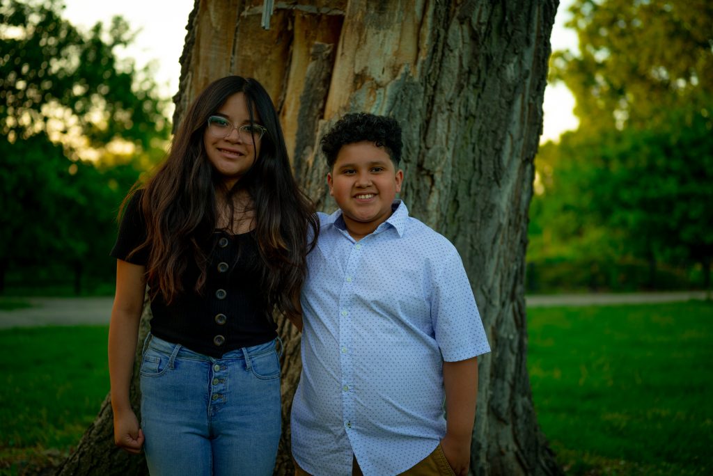 Image: Sophia (right) and Jesse (left) stand in front of a tree in a park, smiling for the camera. Sophia wears jeans and a black top. Jesse wears a patterned button-up shirt and khaki slacks. Photo by Tonal Simmons.