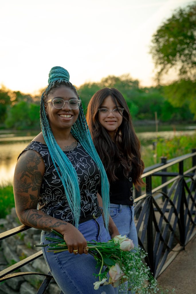 Image: Janet and Sophia lean against an iron fence smiling at the camera. Janet holds a small bouquet of roses and baby's breath. Photo by Tonal Simmons.