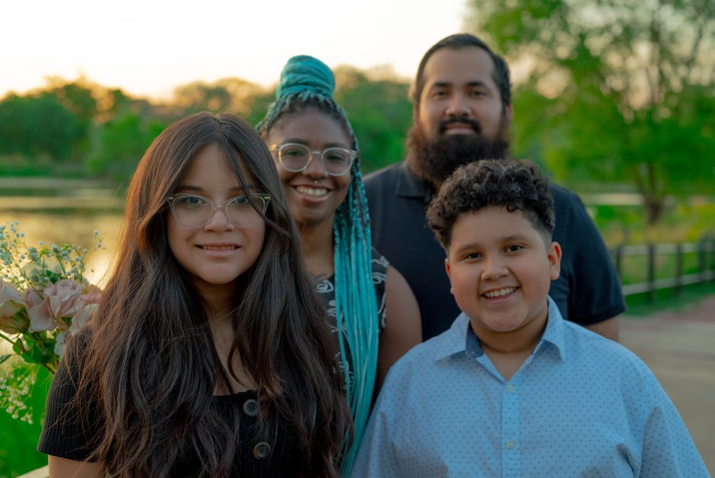 Janet, David, Sophia, and Jesse stand together on a boardwalk in front of a pond in Humboldt Park. Janet and David stand behind Sophia and Jesse, all four are smiling at the camera. Janet wears clear green glasses, has septum and nostril piercings, and wears her hair braided with teal extensions. David has a full beard and mustache, and wears his hair cut close to his head. Sophia wears cat-eye clear glasses. Her hair is long and she smiles at the camera. Jesse has curly hair, wears a patterned button-up shirt and smiles big at the camera. Photo by Tonal Simmons.
