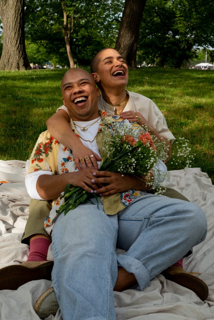 Image: Eli and Lucky laugh together while sitting on an off-white sheet in a field of grass. Eli holds a bouquet of flowers while resting their body against Lucky. Photo by Tonal Simmons.