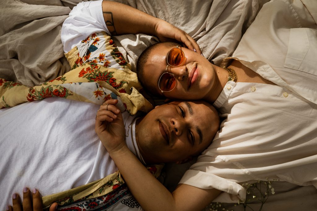 Image: Eli (left) and Lucky (right) lay on an off-white sheet, smiling at the camera. Lucky's right forearm is resting across Eli's chest while Eli's right hand is tenderly cupping Lucky's left ear. Eli is wearing a long-sleeve white shirt under a red, green, and yellow floral short sleeve shirt. Lucky is wearing a white grandfather-collared button shirt with the sleeves rolled up and amber sunglasses. Photo by Tonal Simmons.