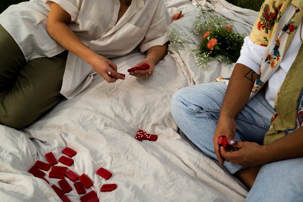 Image: Point-of-View shot of Lucky (left) and Eli's (right) sitting on an off-white sheet playing dominoes. In their hands they have red dominoes. Photo by Tonal Simmons.