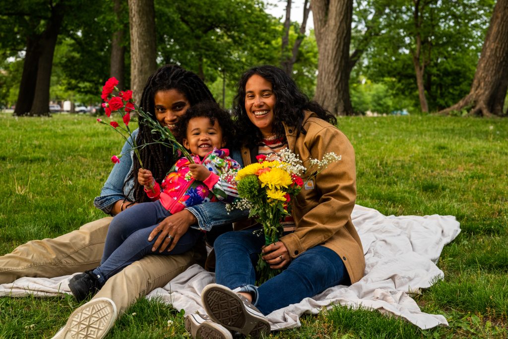 Taryn, Suyan, and Vivi sit on an off-white sheet in a field in Humboldt Park, smiling at the camera. They hold a bouquet of flowers. Photo by Tonal Simmons.
