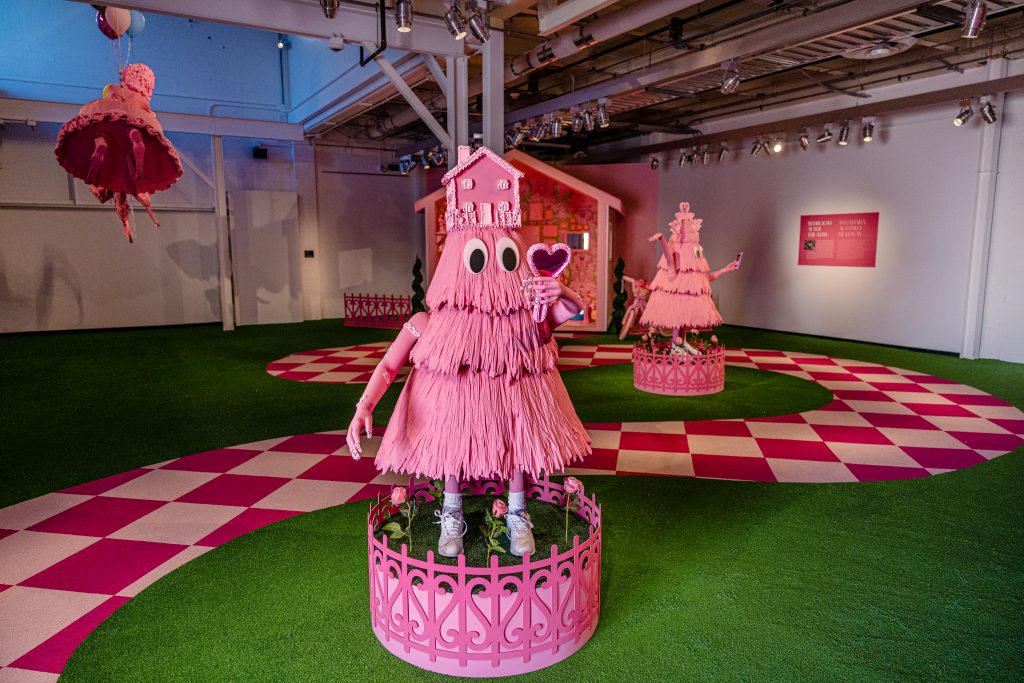 Image: Installation photo of Yvette Mayorga's What a time to be at the Momentary. Centered is a cartoonish pink figure standing on a fenced-in plinth, holding a heart-shaped mirror in one hand. On their head is a small pink house. A pink-and-white checked path cuts through green astroturf behind them. There is a similar figure behind. Image courtesy of Crystal Bridges Museum of American Art.