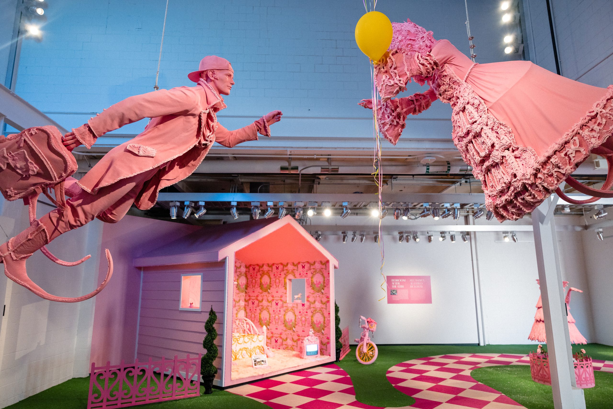 Image: Installation photo of Yvette Mayorga's What a time to be at the Momentary. Two large pink figures hang above a pink and white checked floor with strips of grassy green interspersed. On the floor is a small pink hut with one wall cut away. Image courtesy of Crystal Bridges Museum of American Art.
