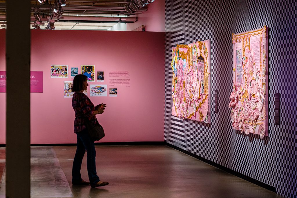 Image: Installation photo of Yvette Mayorga's <i>What a time to be</i> at the Momentary. A museum patron silhouetted against a pastel pink exhibition description. They look at two knit textile art pieces, mostly gold and pink. Some of the material protrudes beyond the canvas. Image courtesy of Crystal Bridges Museum of American Art.