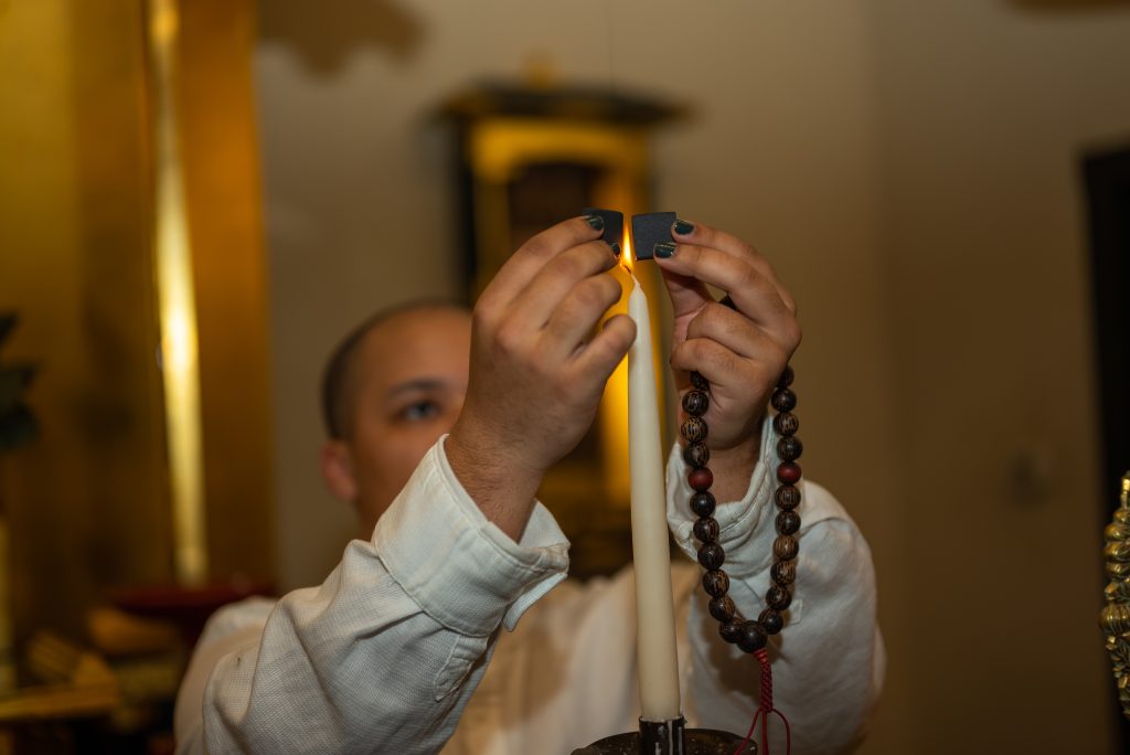 Image: Eli Ryn Brown holding two black chips and raising them around the flame of a burning candle. In their left hand are prayer beads. Photo by Tonal Simmons.