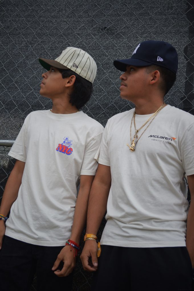 Image: Two brown youth wearing white tee's and baseball hats looking at the stage from the Green Room Area at Miche Fest, 2023. Photo by Luz Magdaleno Flores