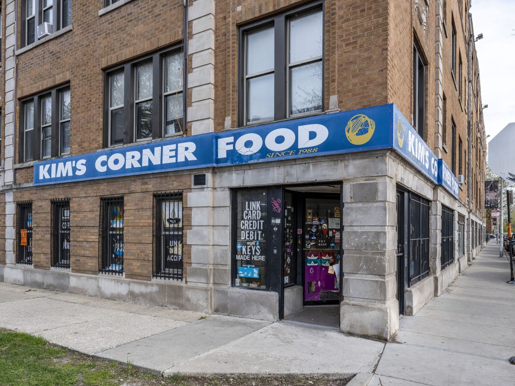 Image: A documentation of the exterior of Kim’s Corner Food, located at 1371 W Estes Ave in Rogers Park. The store is at a corner of a brick and stone, multi-unit residential building. The store’s name, “Kim’s Corner Food,” is in white san serif font, all caps, over a lake blue banner that wraps around the building’s corner. Additional text on the banner reads “Since 1988” with a yellow basket logo. Thomas Kong’s collage works are hung from the inside of the store windows and entrance door; also seen on the store’s vitrine is a list that reads “Link/card/credit/debit/keys/made here.” 2023. Photograph by Guanyu Xu.