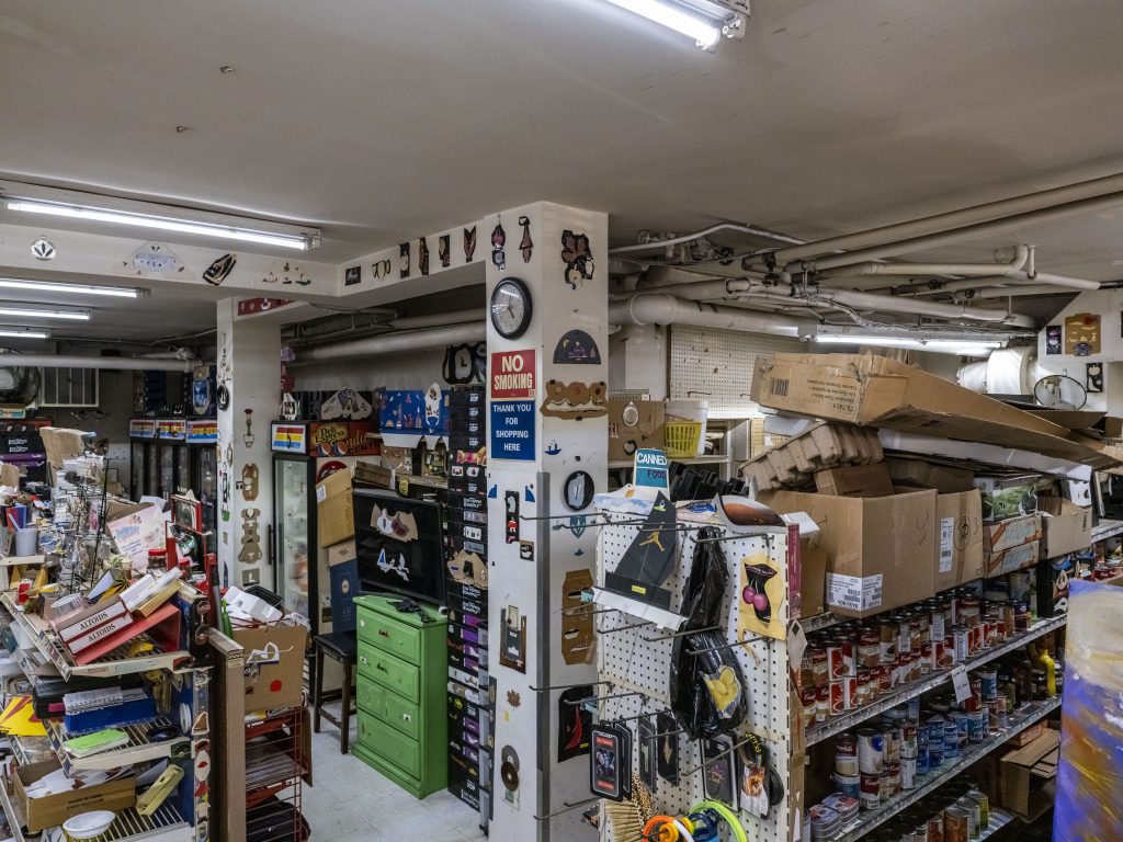 Image: An overview of Kim’s Corner Food’s interior from ground level to show that the store is slightly sunken. The store is packed with products, empty cardboard boxes, and artworks made by Thomas. There are Altoid mints, canned soups and stationary on the shelves. The columns, shown in the middle of the image, are covered with Thomas’s collages among other objects such as a “no smoking” sign and a clock. Between the columns, there is a television sitting on top of a grass green chest, a tall stack of crates. There is also a row of refrigerators further back. 2023. Photograph by Guanyu Xu.