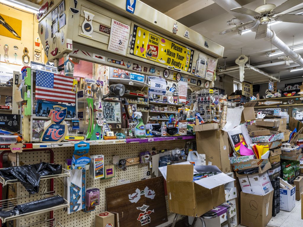 Image: View of the register. The area is swamped by stacks of cardboard boxes that are almost higher than the counter. Miscellaneous small items for sale such as rulers, windshield towelettes, plastic hair clips, and staples are hung beneath the counter. Among all this, artworks of various sizes are hung–many of which have Thomas’s signature sticker label “be happy” on them. The counter is crowded by all sorts of products, from lighters for sale to faded photographs, a desk lamp, packs of cigarettes, hand sanitizer, and more artworks decorating the display shelves. In the background, a cigarette shelf is mostly empty. Above the very worn-out shelves, a yellow metal sign reads “please have your ID ready.” Further to the right, a giant piece of plastic in the shape of a key is suspended from the ceiling. A ceiling fan is also seen at the upper right corner of the photo. There isn’t a single empty place not decorated with some kind of collage artwork. 2023. Photograph by Guanyu Xu.