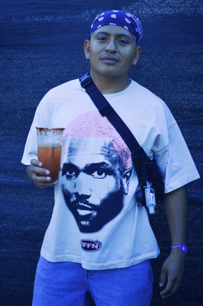 Image: A brown person wearing a blue bandana and white shirt with a printed face on it holding a Michelada at Miche Fest, 2023. Photo by Luz Magdaleno Flores