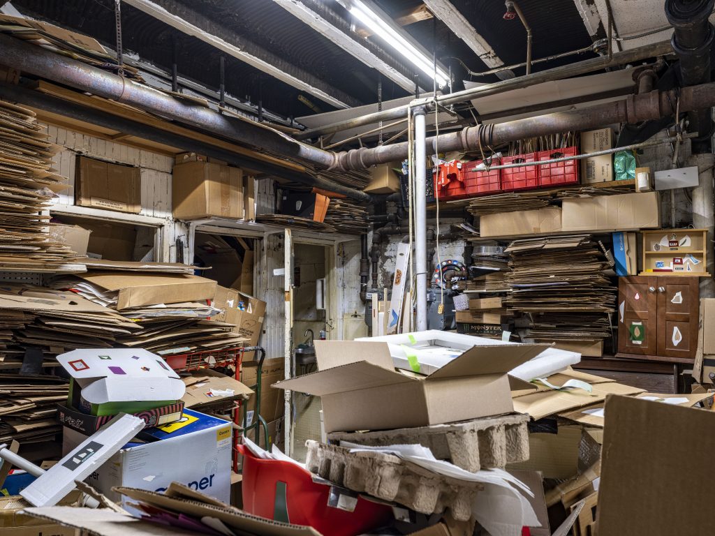 Image: A documentation of the storage area in the back of the store. Between 2015 and 2019 the experimental project space, “The Back Room,” was organized here. Stacks of cardboards–almost floor to ceiling–can be seen at the left and in the far back; they are all Thomas’s artworks. At the front, there are more stacks of cardboard boxes, pulp molded trays, a red helmet, and foams. In the back to the right, some cabinets and shelves have Thomas’s collages stuck to the surface. Old ceiling pipes can be seen at the top of the image. Three doorways are barely visible behind the cardboard stacks. 2023. Photograph by Guanyu Xu.