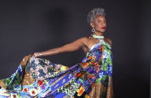 Cynthia Oliver is photographed wearing a colorful, multi-fabric sleeveless dress, red lipstick, and dangling earrings. Oliver is a tall Black woman with short gray hair. Her right arm is stretched behind her holding up her dress. The background is a neutral charcoal-black color. Photo by LaTosha Pointer, courtesy of Cynthia Oliver.