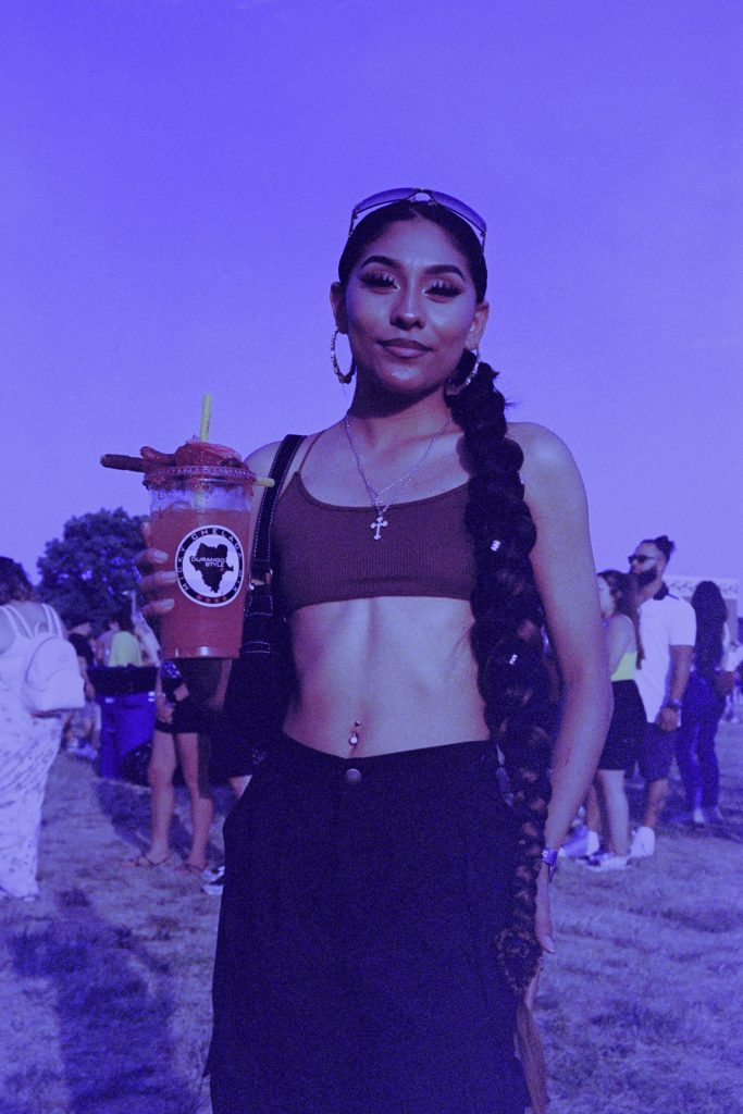 Image: A brown person wearing glasses on their head, a brown bralette, with a gold cross necklace, shiny belly button ring, and black pants holding a Michelada at Miche Fest, 2023. Photo by Luz Magdaleno Flores