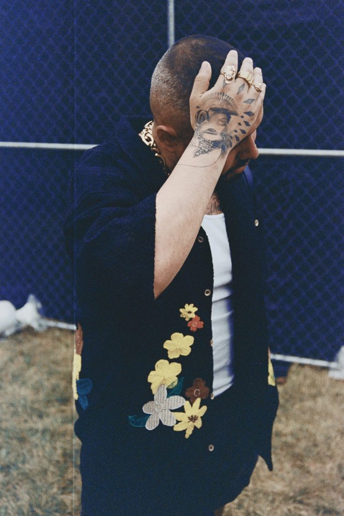 Image: A tattooed person wearing gold jewelry, a short sleeve button up with flowers embroidered, with a peeking white daygo posing during Miche Fest, 2023. Photo by Luz Magdaleno Flores