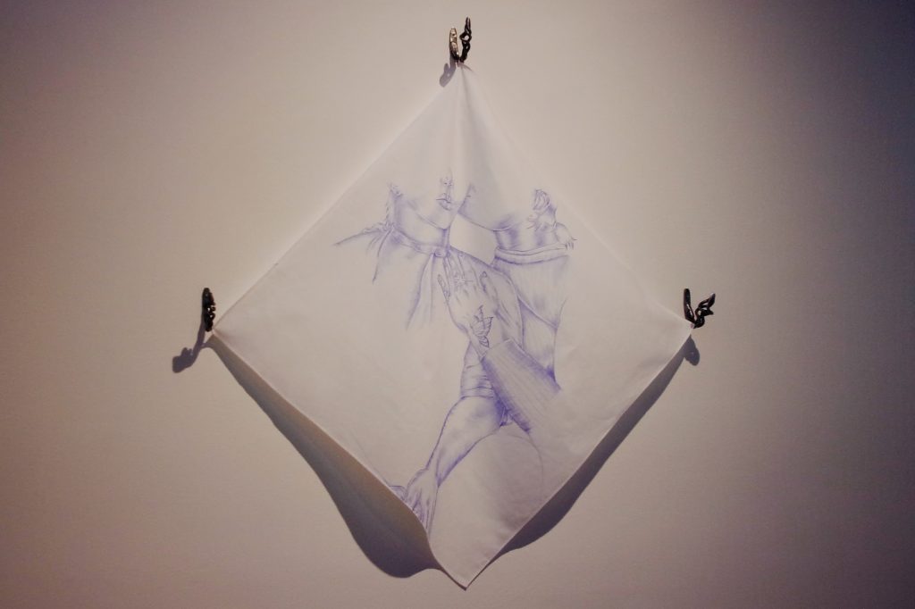 Image: Cariñito by Vani Aguilar. A white bandana pinned up to the wall, with a blue ballpoint pen drawing of a person kissing another on the cheek. Photo by Luz Magdaleno Flores.
