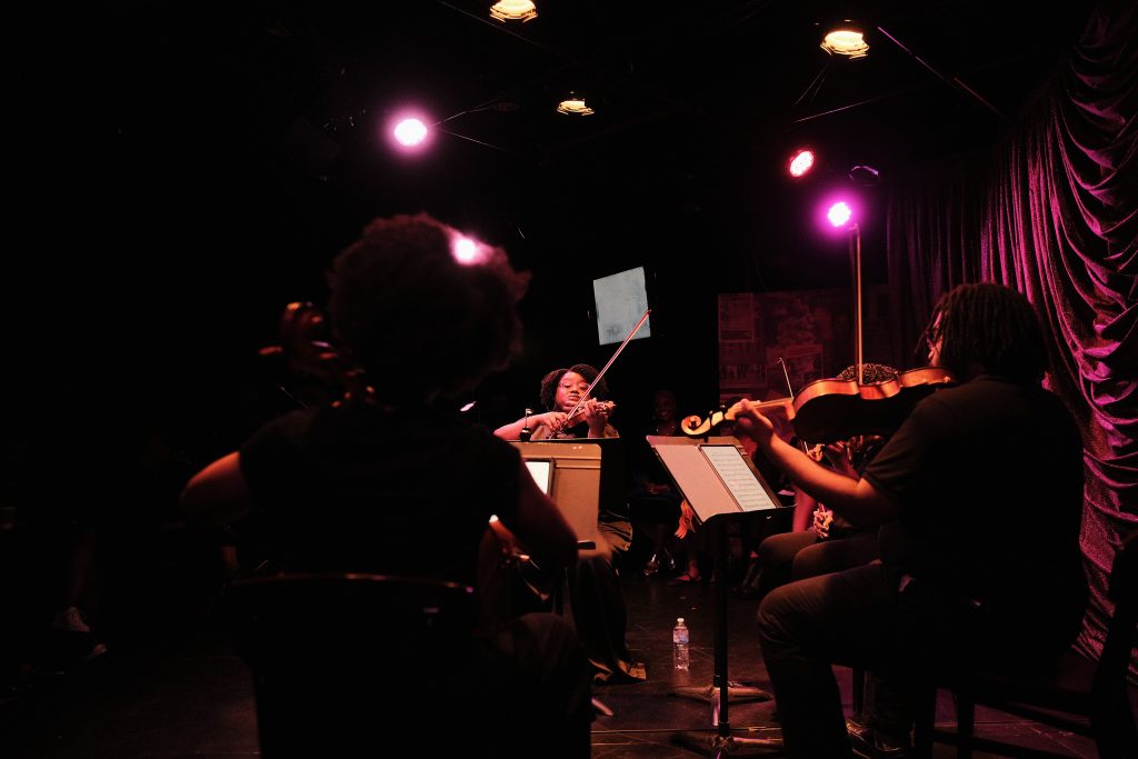 Image: D-Composed's string quartet performs an arrangement. One of the musicians is shadowed, their back facing the camera, while the other three are visible, illuminated by soft purple light as they play their instruments onstage. Image credit: Sulyiman Stokes.