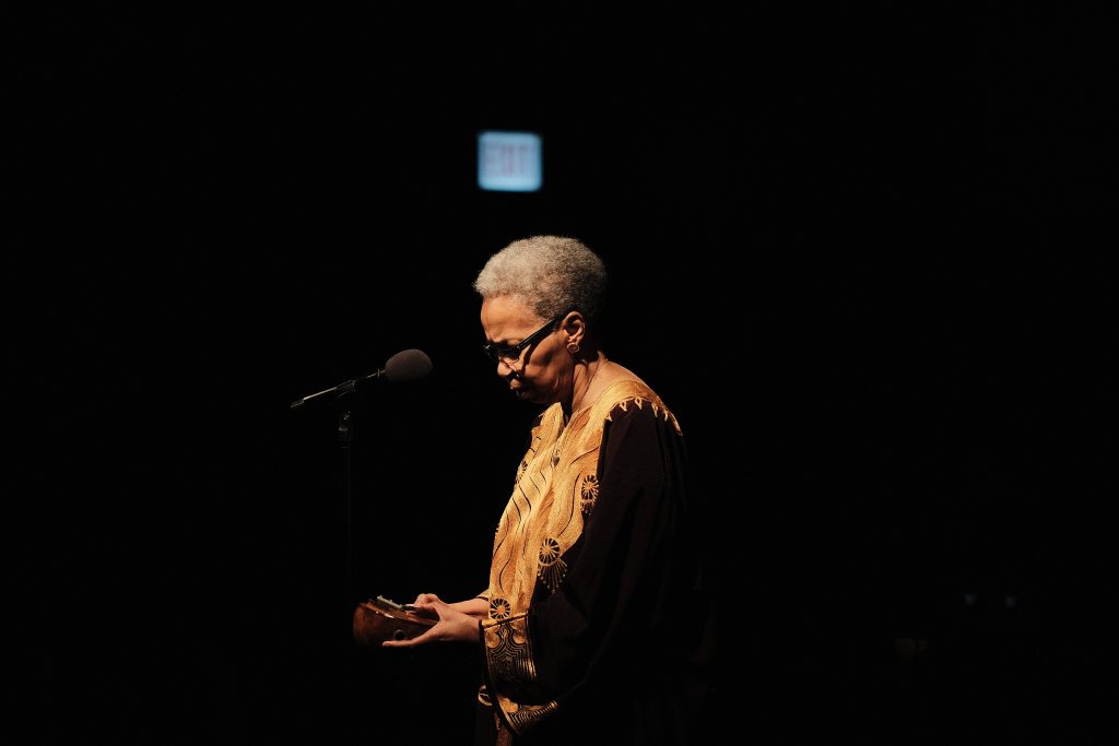 Image: Storyteller and instrumentalist Shanta Nurullah, a Black woman with short, gray hair, stands in front of a microphone with her gaze focused on the instrument clasped in her hands. She is contrasted against a black background. Image Credit: Sulyiman Stokes.