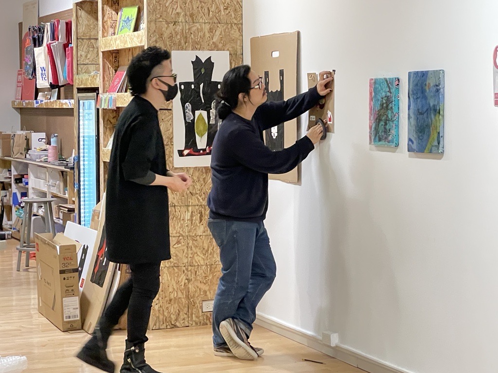 Image: Greg Bae stands at a white gallery wall pinning a collage by Thomas Kong with a student observing behind him. Two other collages by Thomas hang in the background. The pieces hung are small to medium, square pieces of cardboard with indistinguishable cut out images pasted to it. Small abstract paintings by Yae Jee Min can be seen to the right of Thomas’s works. Photograph by Cathay Hsiao.