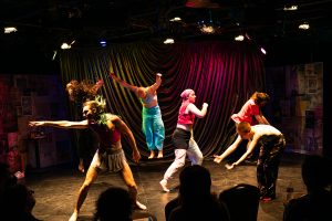 All six performers are photographed in motion against a multicolored curtain: in the foreground, a performer is standing with their leg spread apart, reaching out towards the left side of the stage. Behind them, another performer is barely visible. They’re mid jump with their arms thrown backwards. Beside them, a dancer in electric blue pants is hovering, mid jump, their arms stretched above them in a Y shape. Another dancing wearing a bandana appears to be stepping towards the right side of the stage while their arm is bent and held in front of their chest. The remaining two performers are standing in opposing directions on the right side of the stage, their knees slightly bent, while they lean forward and stretch their arms toward the ground.