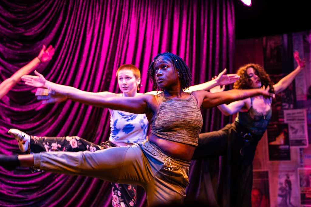 Image: QDF company lead A'Keisha Lee and two other QDF company members perform onstage at Steppenwolf Theatre. A Black dancer with short locs is centerstage, wearing brown satin pants and a brown tank top. They're looking to their right while outstretching their arms and lifting their right leg so that it's horizontal. Behind them, two white dancers mirror this position. One has orange hair and is wearing floral pants and a blue and white tank top. The other has long, curly brown hair that falls in their face and they're wearing a lacy bodysuit. Image credit: Kitty Phillips. 
