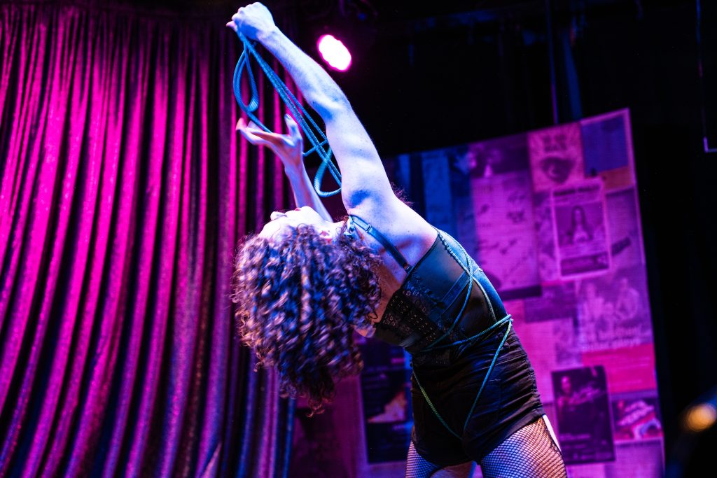 Image: QDF company member Erobus Lane Clementine performs onstage on Steppenwolf Theatre. A white dancer arches backwards, framed between a magenta curtain and a wall of posters. Their head is horizontal as they look up at a blue rope they're pulling upwards from their body. Their long, curly brown hair hangs below their head. They're wearing a bodysuit and shorts that reveal their fishnet tights. Image credit: Kitty Phillips. 