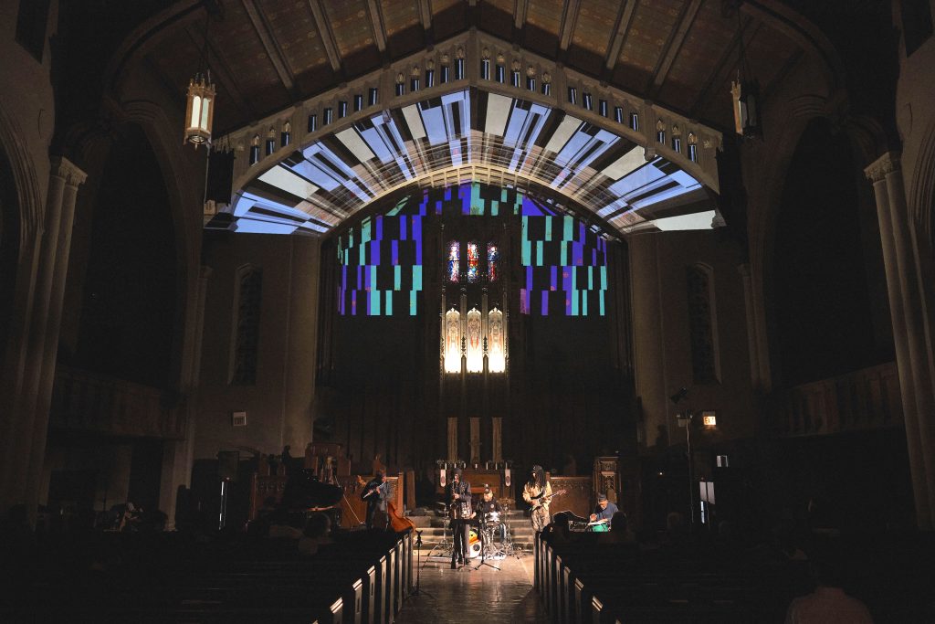 Image: The opening ceremony for Max Li's exhibition. Band Xochipilli and a light show by Maddie Ma on First Presbyterian Church's stage. Colorful stained glass art and lights are behind the stage, while a colorfully lit arch shines over the stage. People are watching the show from the church pews. Photo by Sulyiman Stokes.
