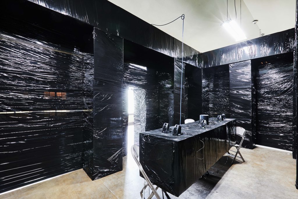 Image: Installation view of Twenty-Four Thinking Positions, multi-media, Max Li, 2023. The tables and walls of the church are covered in black plastic bags from floor to ceiling. There are speakers along the table and three metal folding chairs around it. Natural light flows strongly through the entryway of the exhibition. Image courtesy of Max Li.