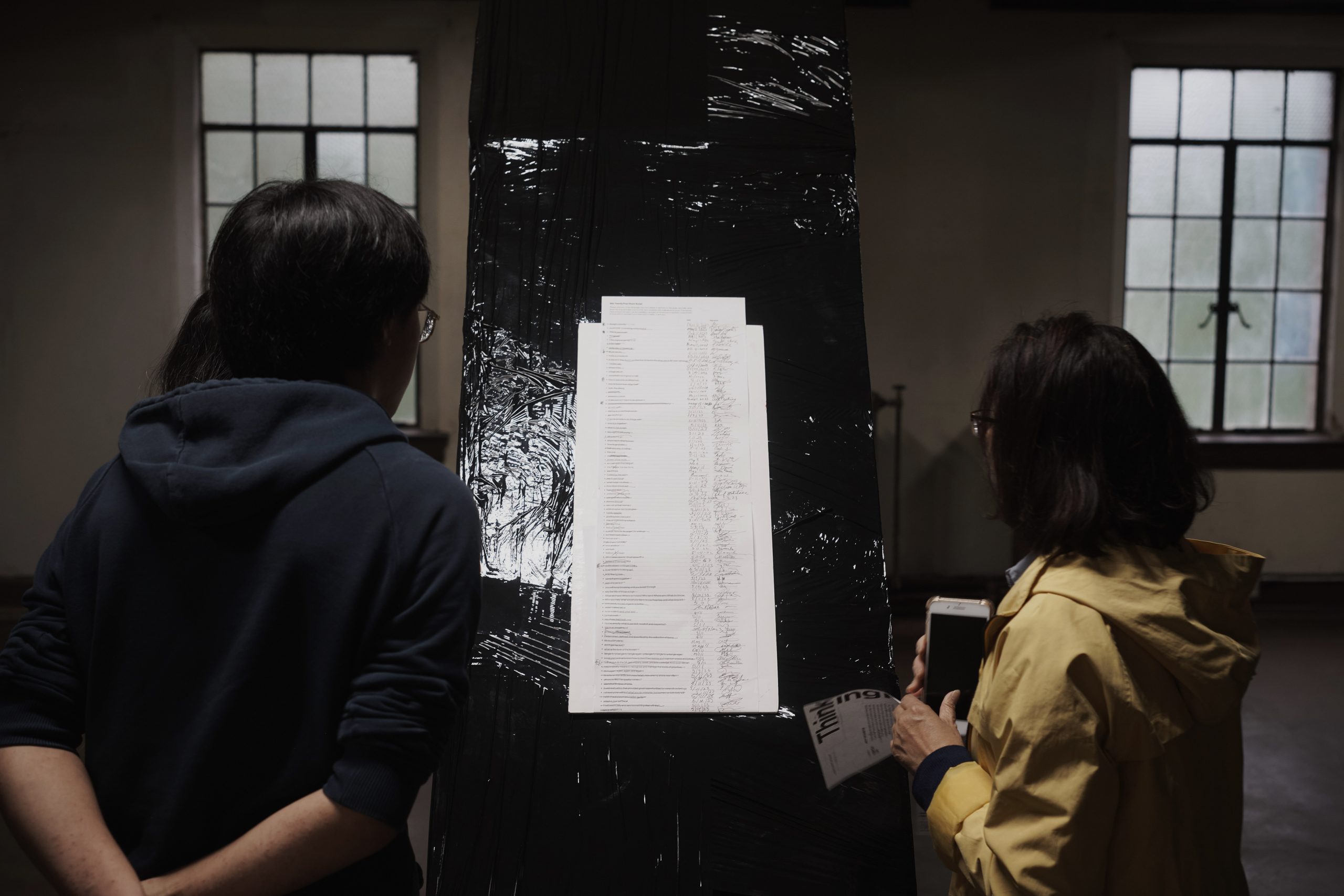 Image: Two people both wearing sweaters silently read from a list of phrases written by Max Li for his installation at First Presbyterian Church. The list is on a long white sheet of paper. It is laid on top of a column which is wrapped in black plastic bags. The column is in between two of the church's windows. Photo by Sulyiman Stokes.