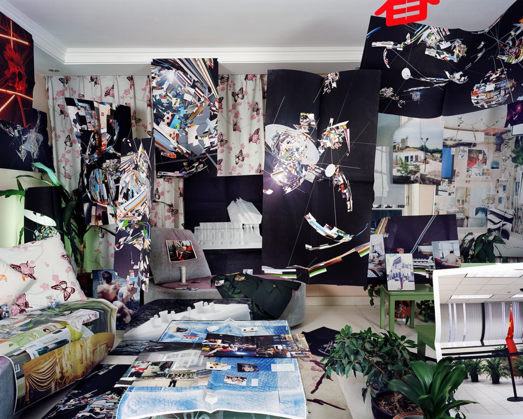 Image: Guanyu Xu, Parents’ Bedroom, 2018. Archival Pigment Print. A photo of a room with dark walls. Printed photos are displayed and laid all over the room and across the walls. Courtesy of the artist.