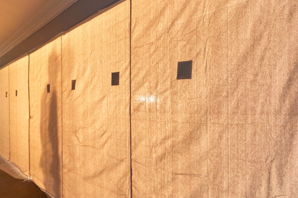 Image: Installation view of 24 Thinking Positions, picturing the backside of walls which consist of sections of orange-tan scrim about 3.5-feet wide with 1-2 tintypes per section. The tintypes are positioned about five feet from the ground. Behind the scrim we can see the silhouette of a visitor. Image courtesy of Max Li.