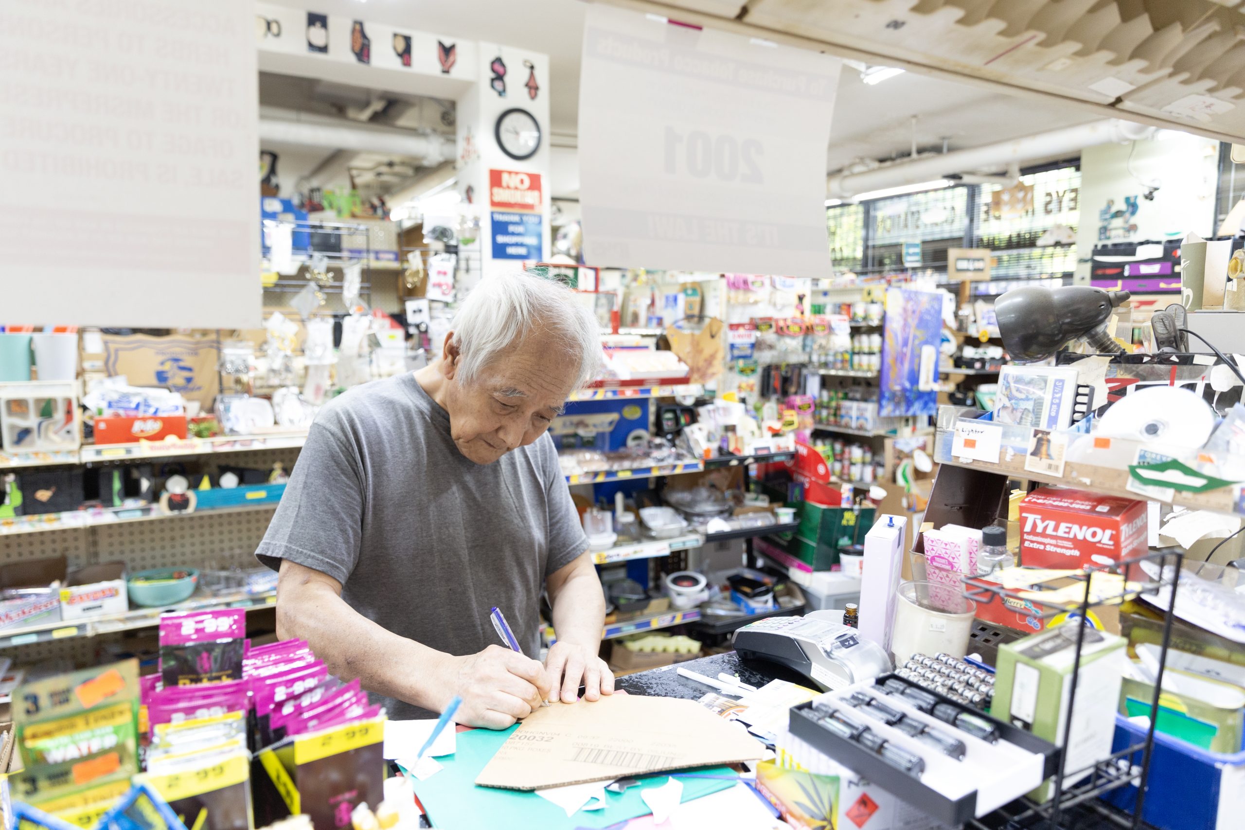 Image: Thomas Kong, an elderly, gray-haired man in a gray T-shirt is seen working behind a store counter. He is seen writing something on a piece of cut cardboard. Photograph by S.Y. Lim